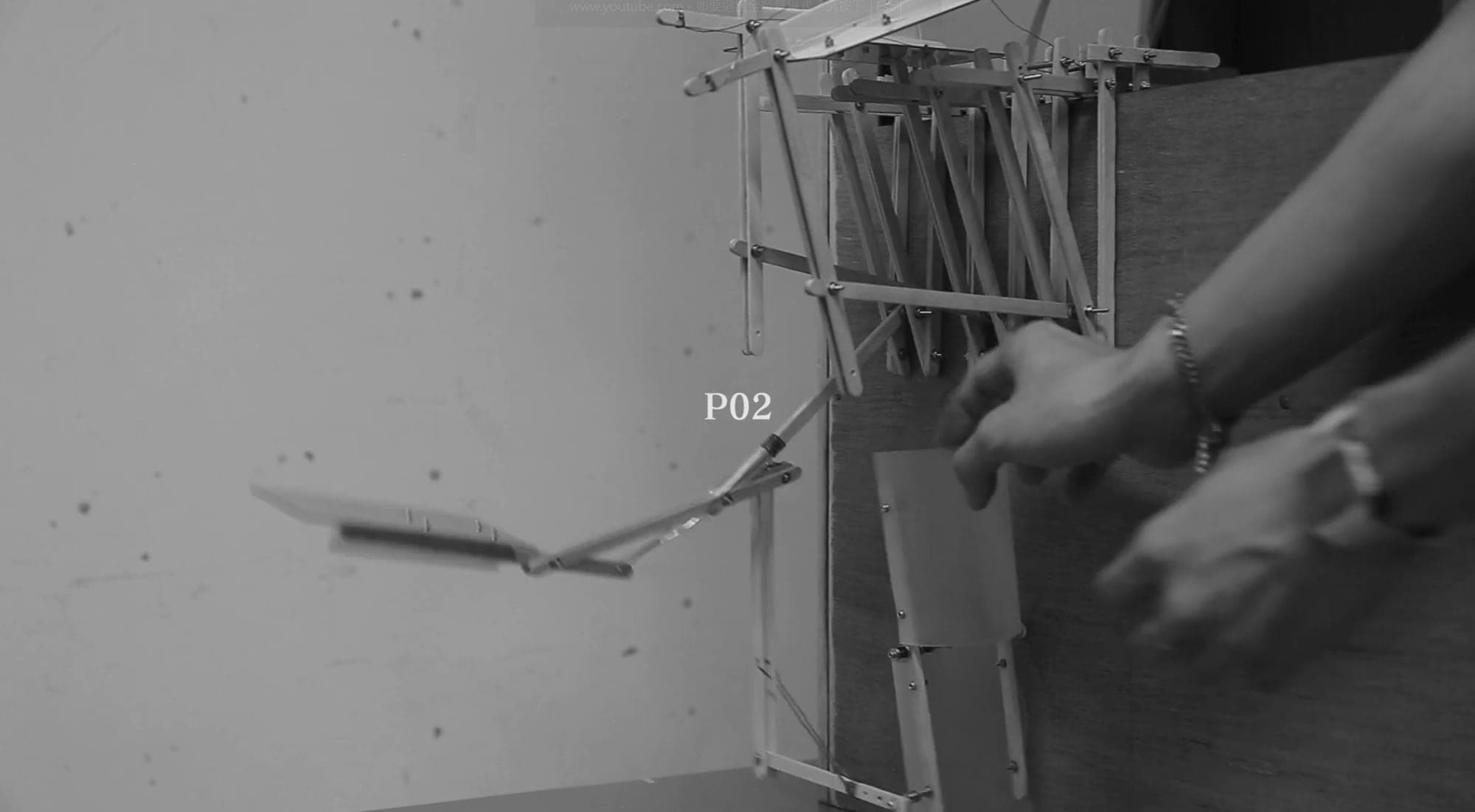 Video still — hands operating parasite P02, a scissor arm swinging a tray out from the host wall