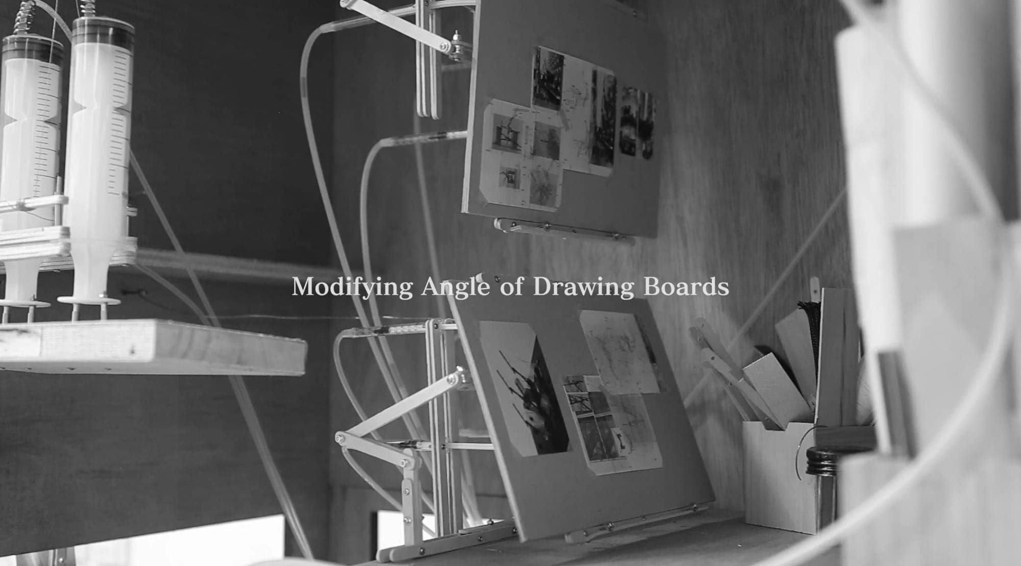Video still — drawing boards tilting on jointed arms as the parasite modifies their angle