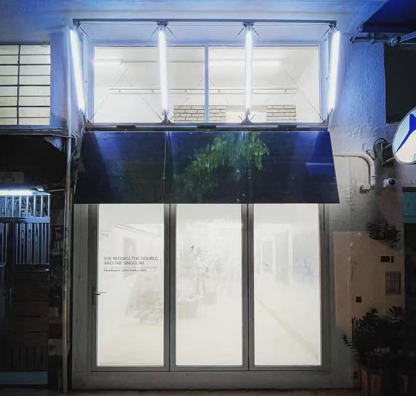 WOAW Gallery at night — the folding steel awning lifted above the glazed storefront, upper window glowing, exhibition titles printed on the glass